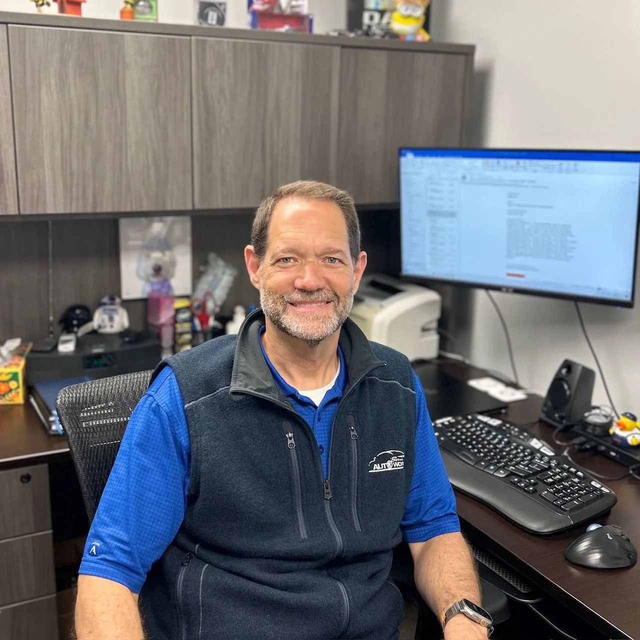 Man smiling at his desk in an office, with a monitor, keyboard, and cabinet behind him.