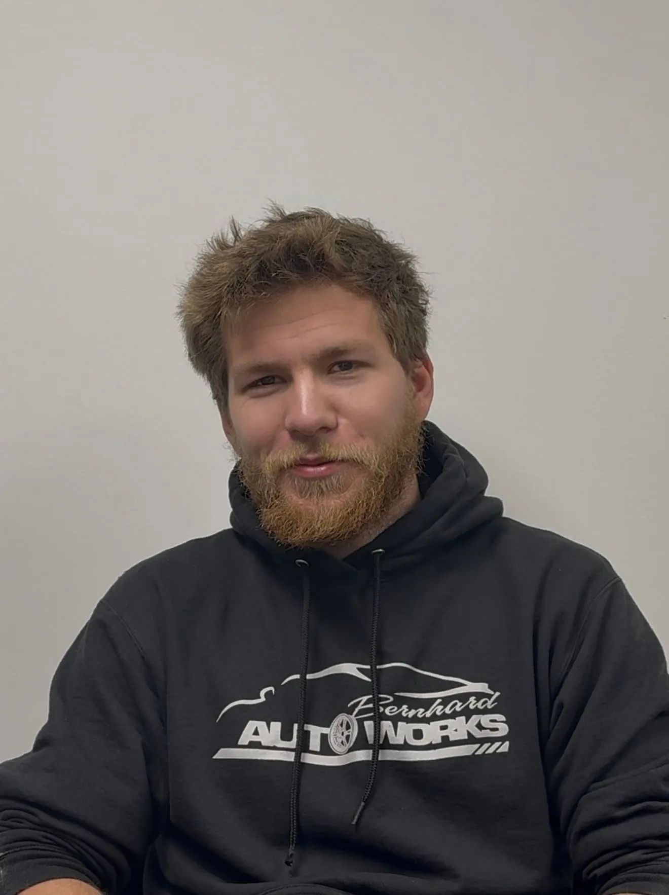 Person in a dark hoodie with a beard, seated against a plain wall, looking at the camera.