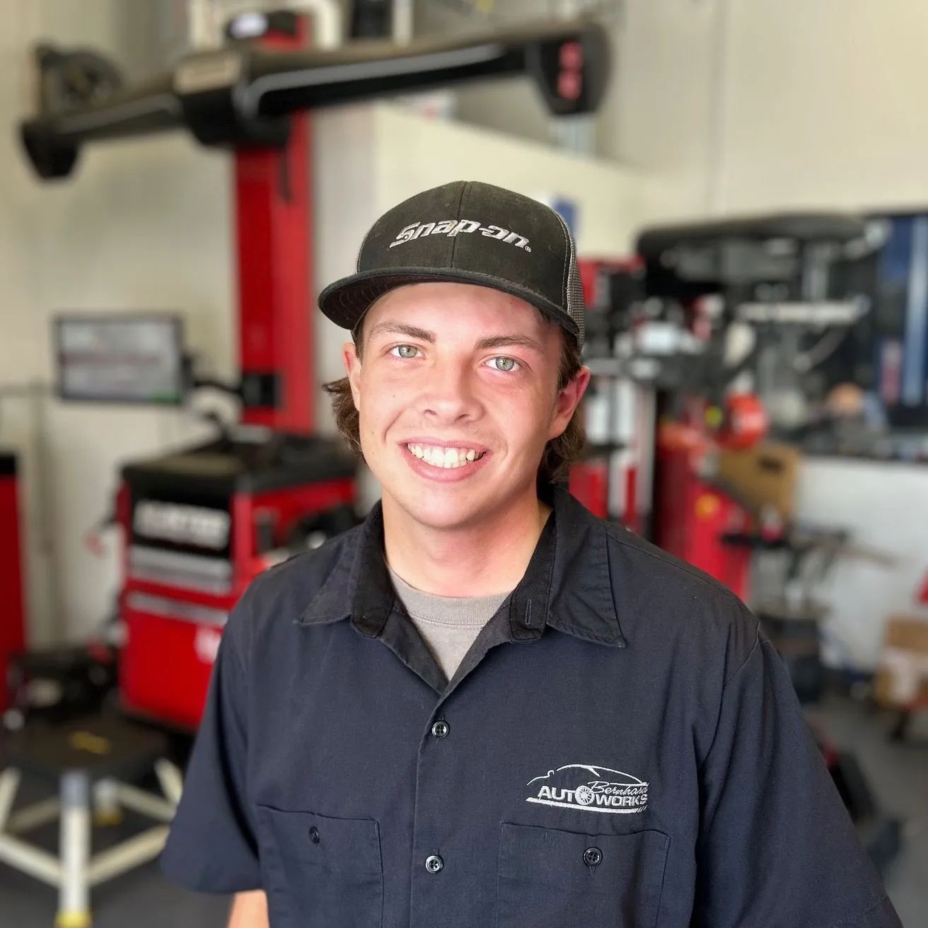 Smiling mechanic in dark shirt and cap standing in an auto repair shop with red lifts and equipment.