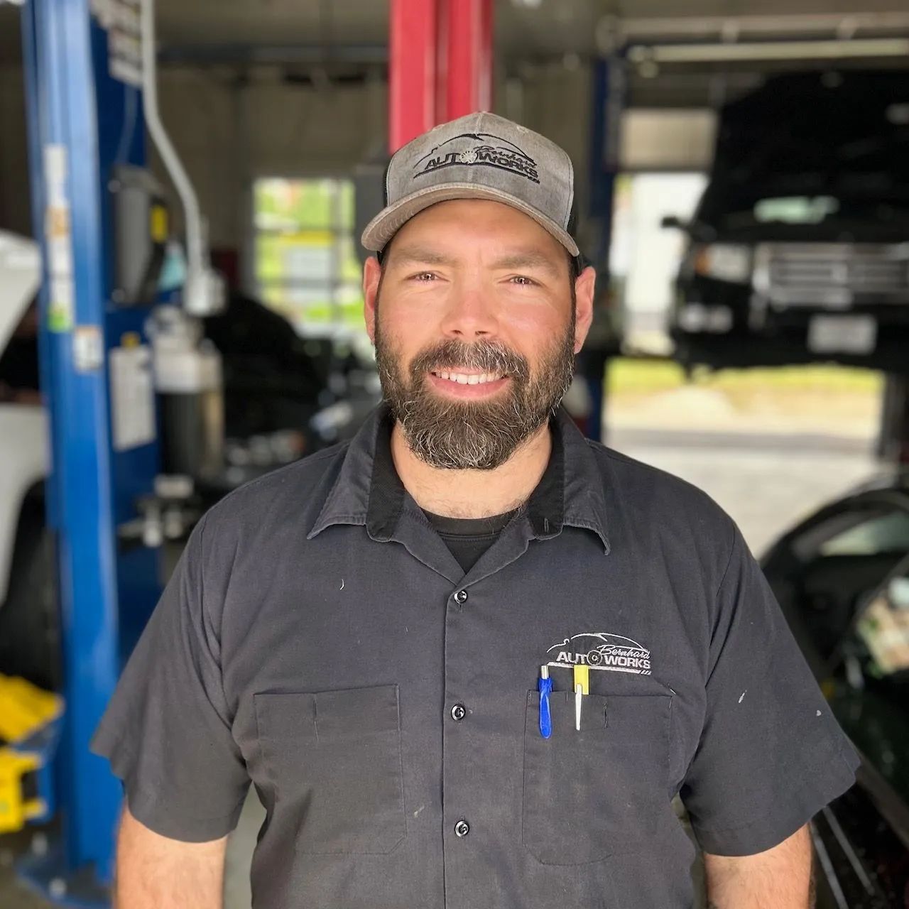 Mechanic in black shirt and gray cap smiling in an auto shop with car lift behind him