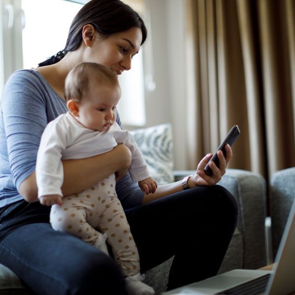 Woman holding a baby on a couch, looking at a smartphone beside a laptop.
