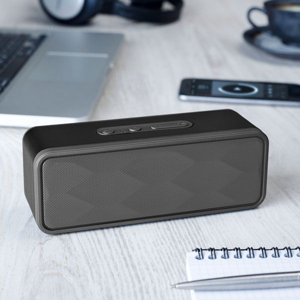 Black portable Bluetooth speaker on a desk with a laptop, notebook, and remote in the background