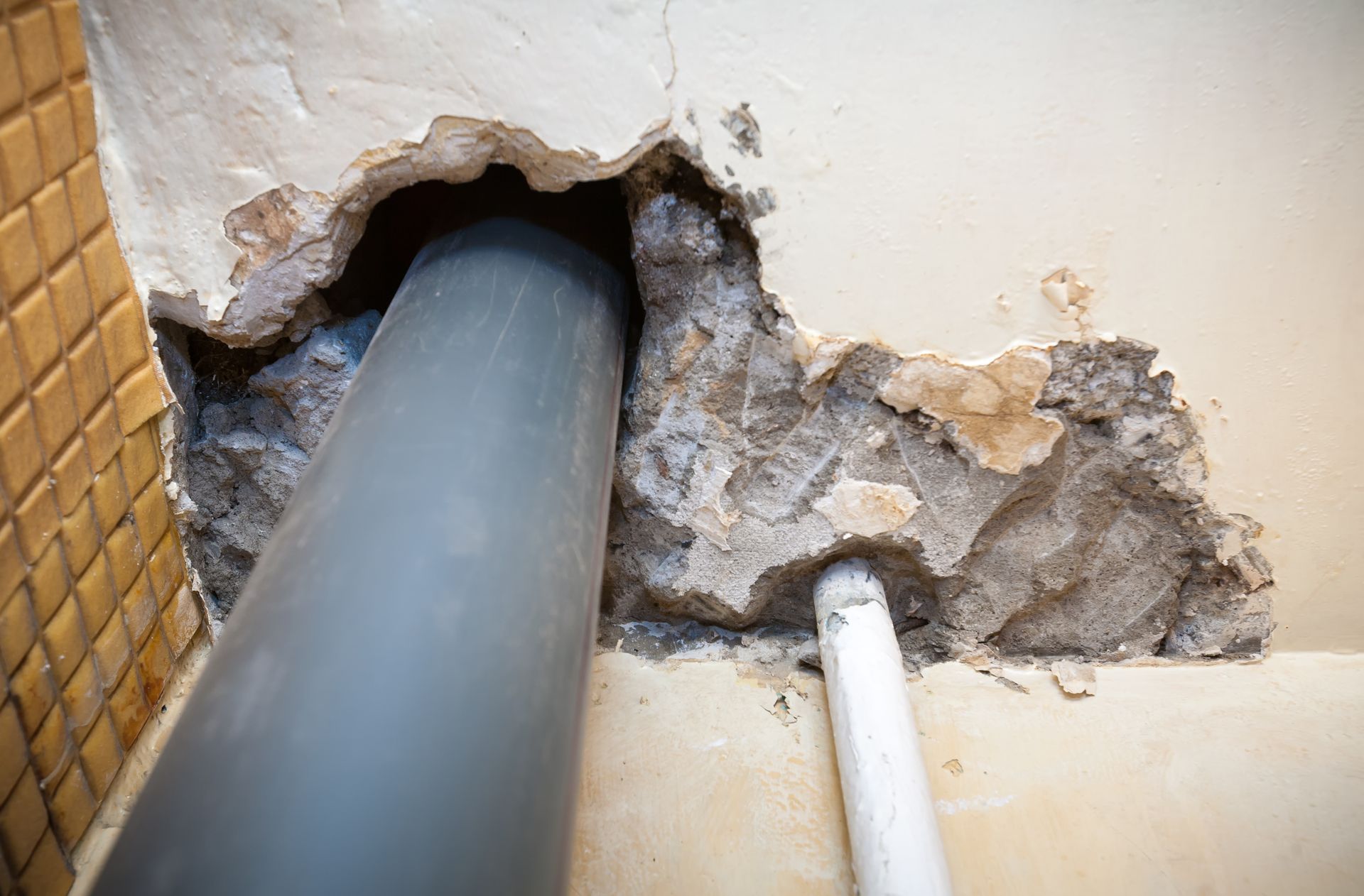 Damaged wall with exposed gray pipe and cracked plaster near a white pipe.
