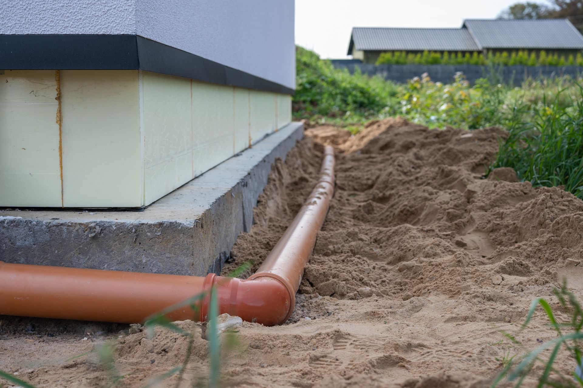 Orange drainage pipe along a house foundation beside a sandy trench and grassy yard