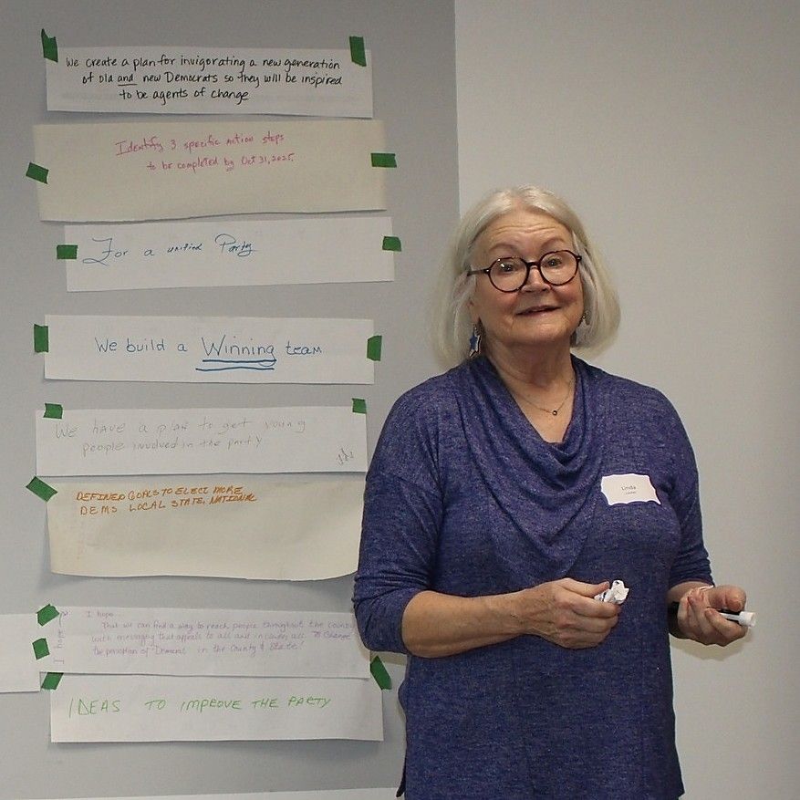Woman with glasses standing by a wall with notes; she holds a marker and smiles.