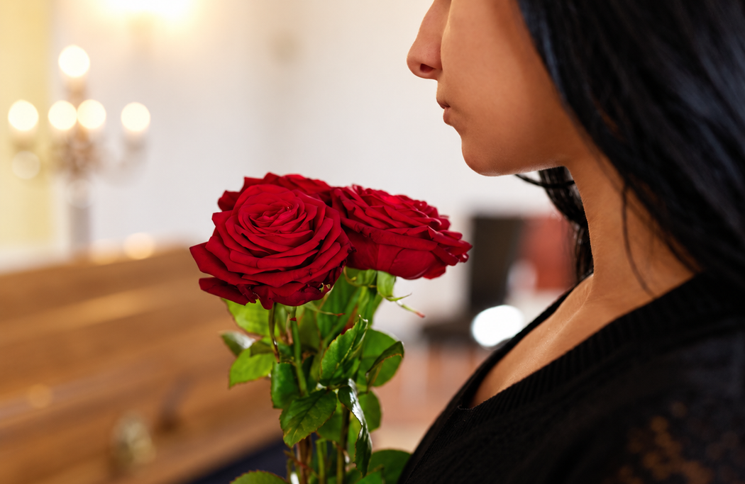 Una donna vestita di nero tiene in mano delle rose rosse accanto a una bara di legno in una cappella illuminata da una luce soffusa.