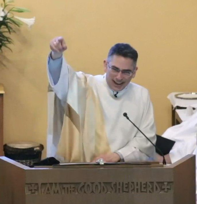 Priest in white robe speaks at a church lectern, raising one hand during a sermon.