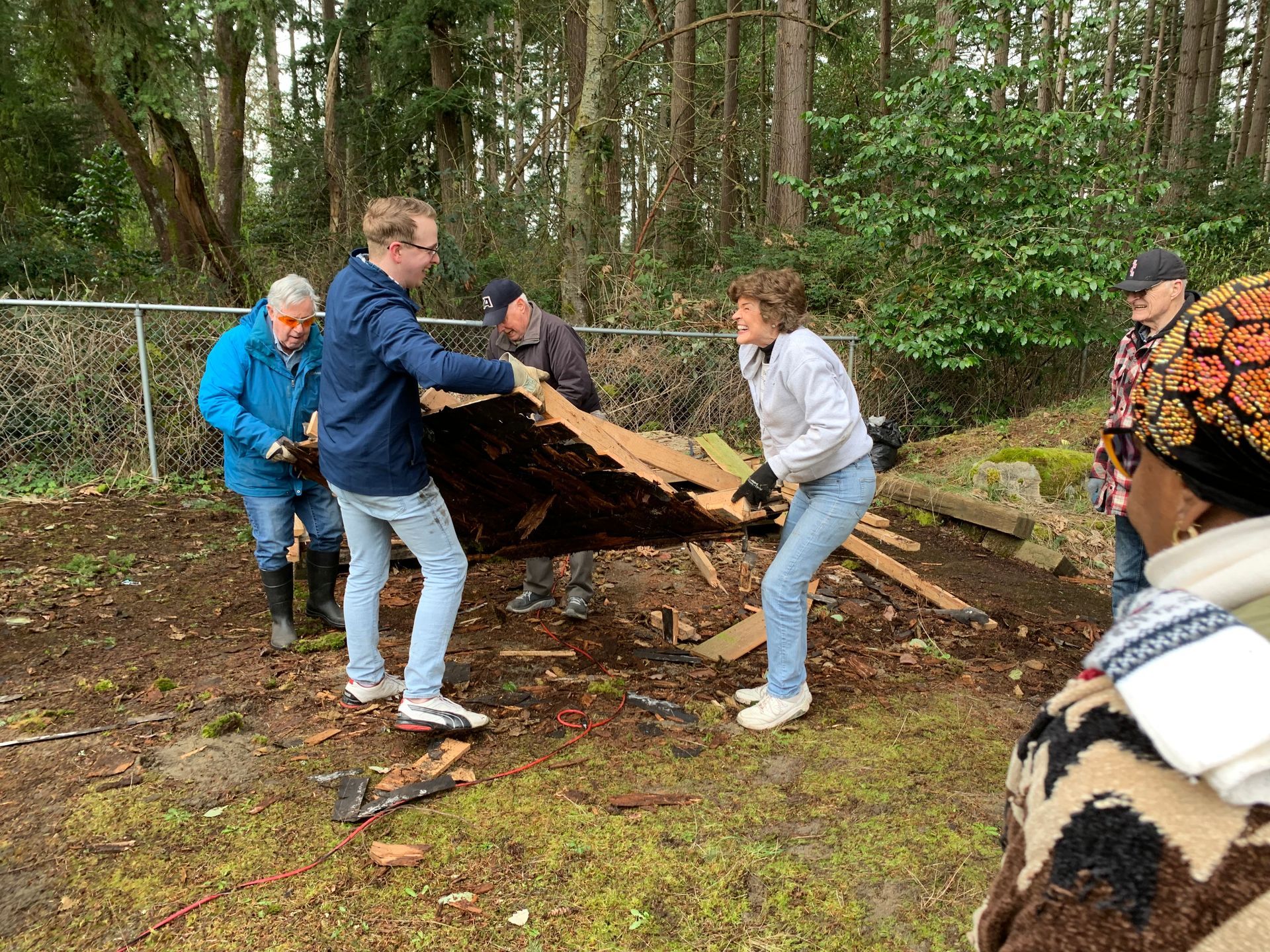 Four people lift and carry a large fallen tree trunk in a wooded area.