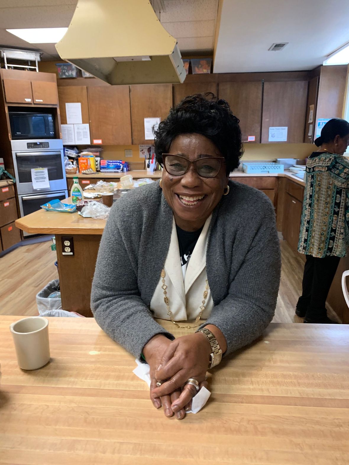 Woman smiling at a kitchen table in a cozy break room, with cabinets and supplies behind her.