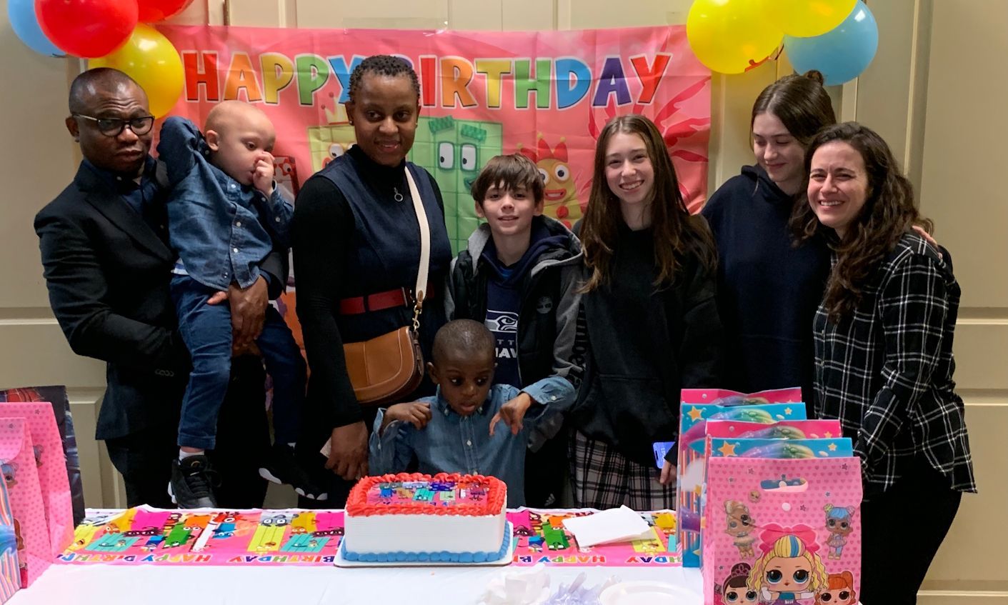 Birthday group posing behind a cake and colorful balloons, smiling at a decorated table.