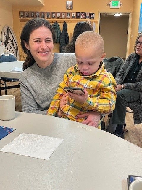 Smiling adult holding a child in a yellow plaid shirt at a table in a community room