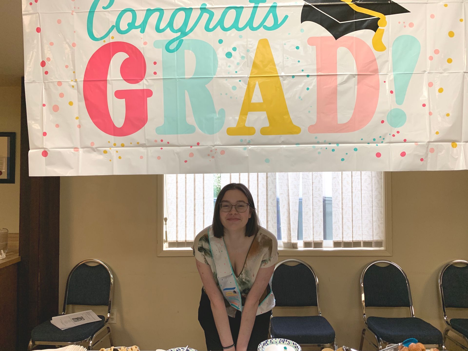 Person standing under a colorful “Congrats Grad!” banner in a decorated room