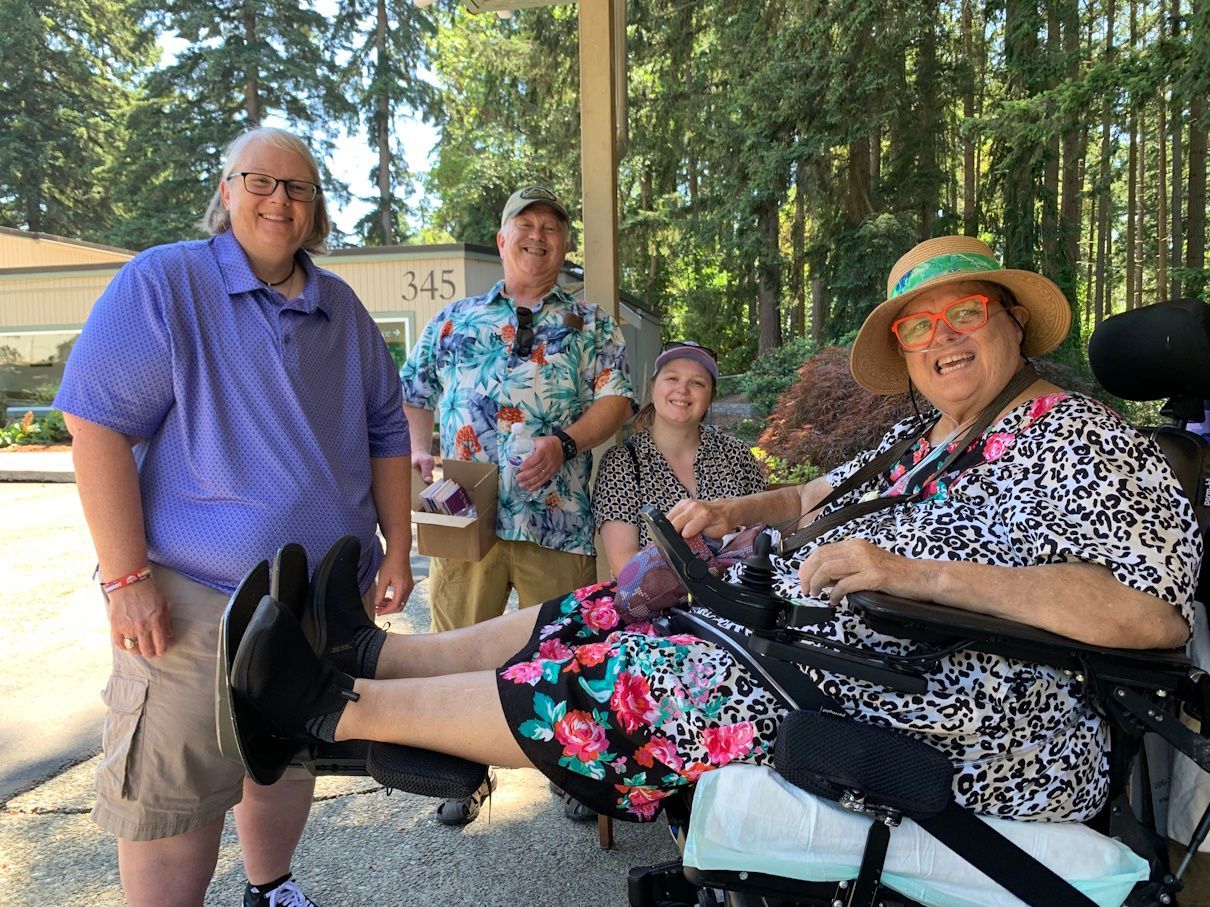 Four people sitting and standing outdoors in a sunny wooded area, smiling at the camera.