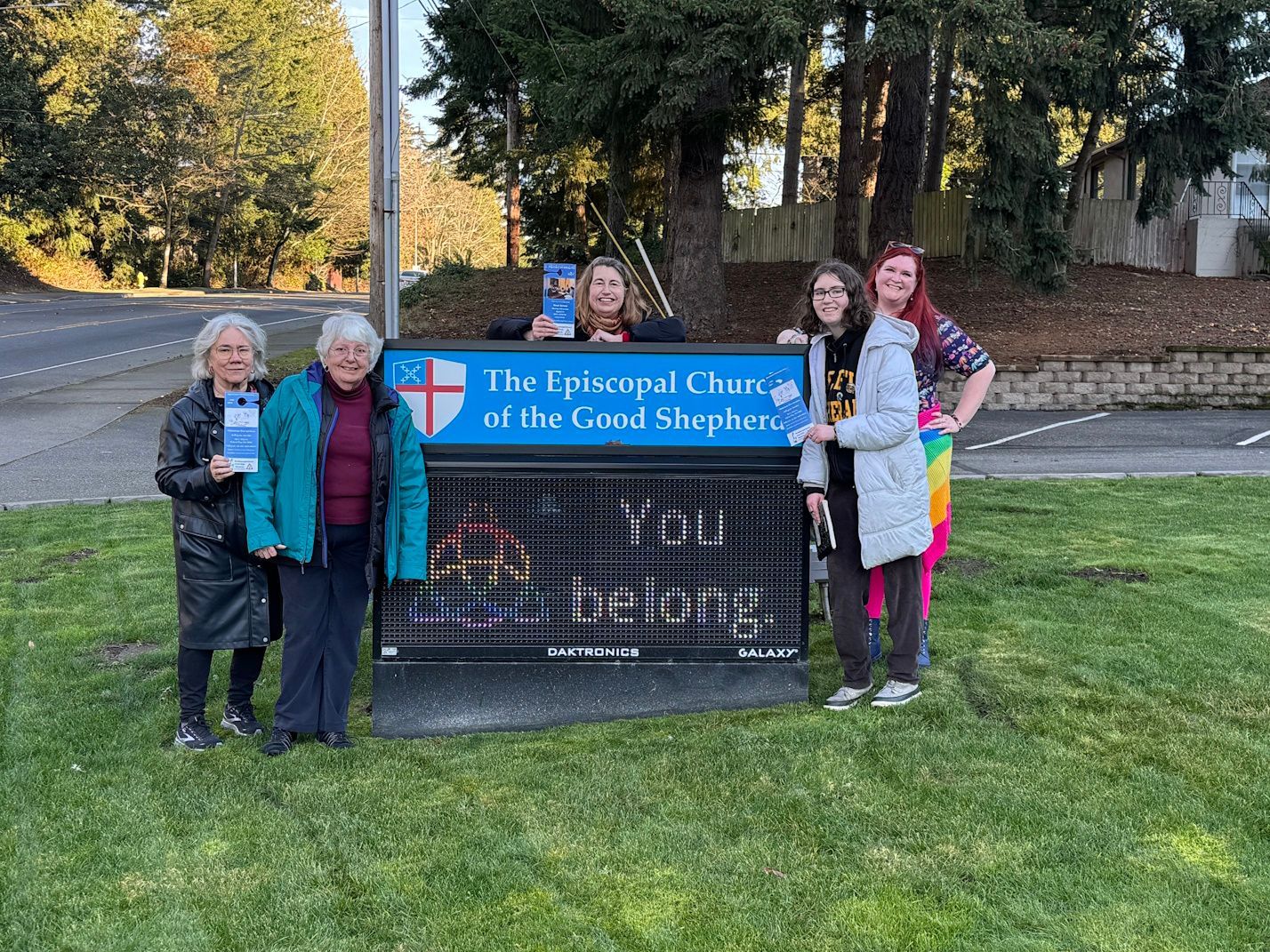 Four people stand beside a sign for The Episcopal Church of the Good Shepherd outdoors on a grassy lawn.