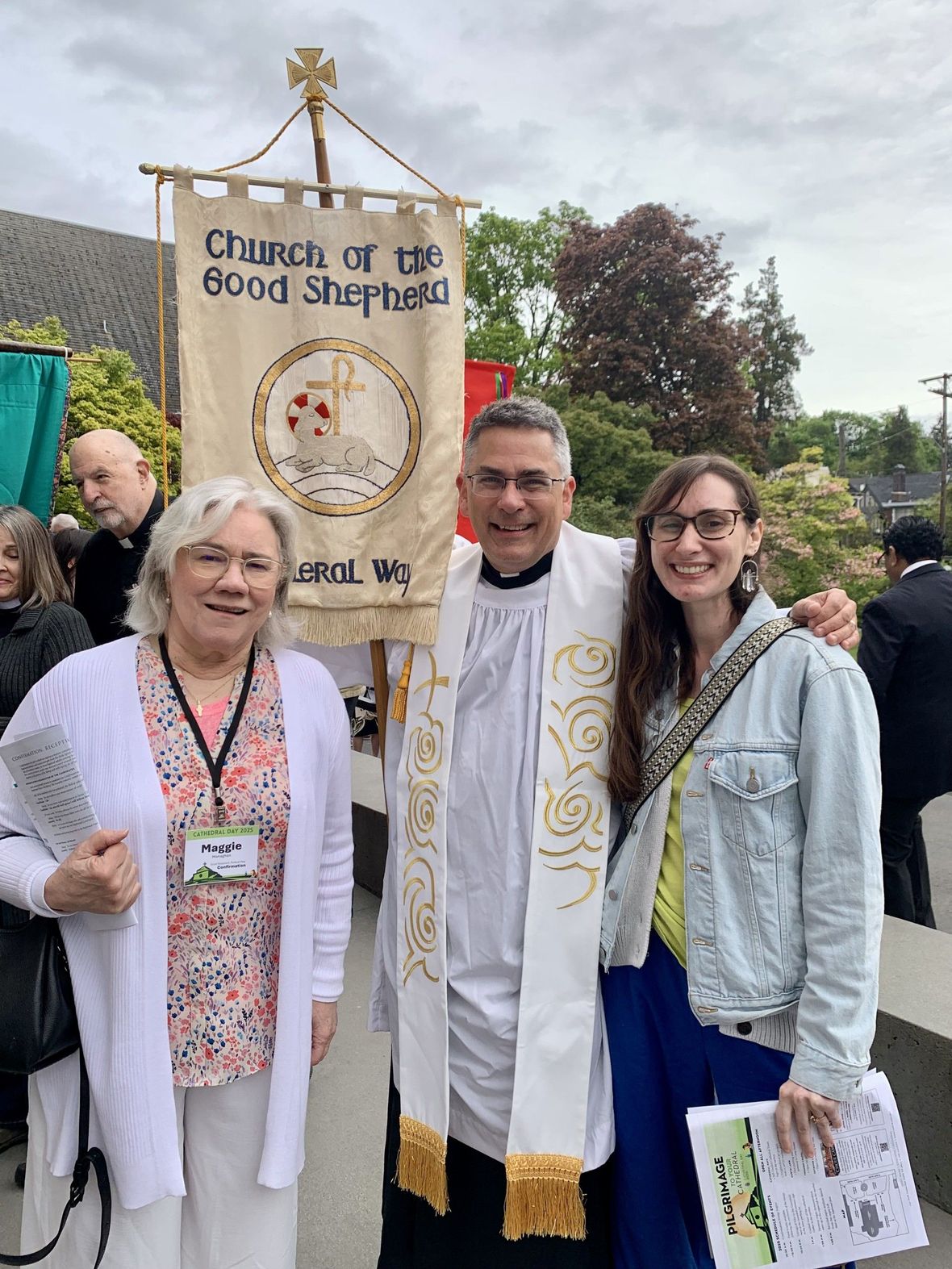 Three people in white stoles stand by a church banner outdoors at a service rally.
