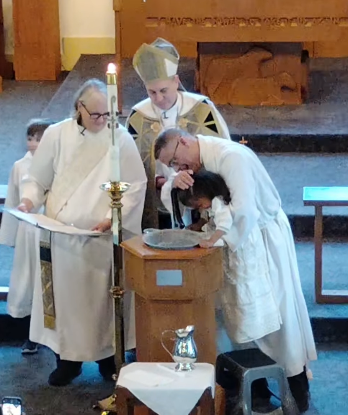 Clergy in white robes performing a baptism at a church font, with one person leaning over a child.