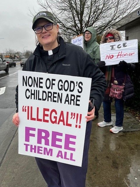 Protester holding sign reading “None of God’s children are ‘illegal’!! Free them all” at a rainy roadside rally