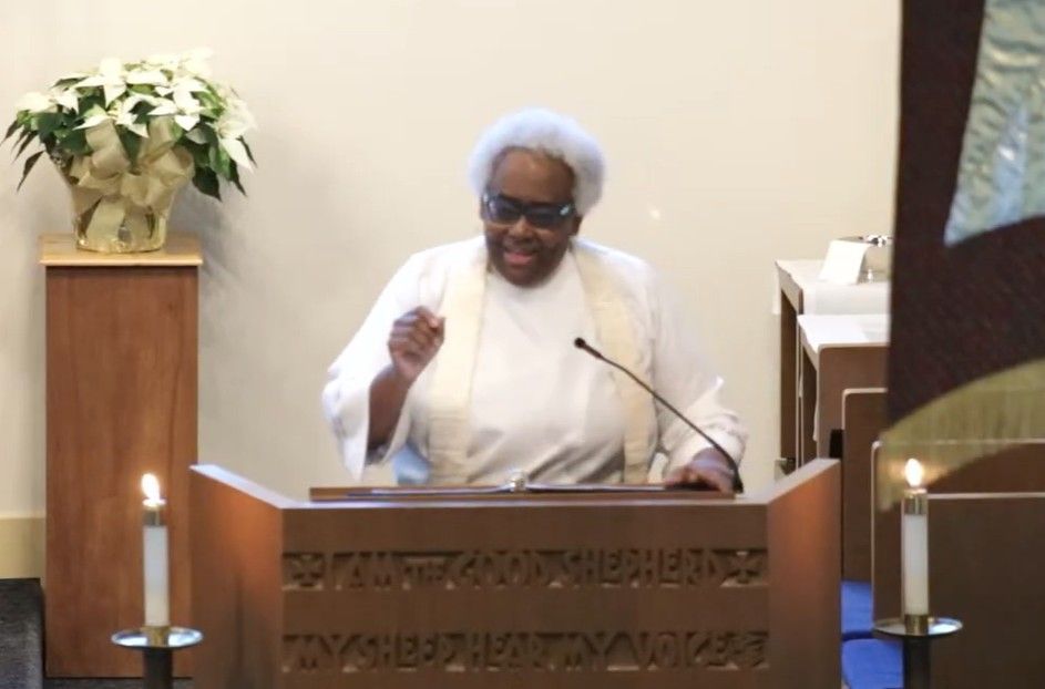 Person speaking at a pulpit in a church, wearing white with a microphone and candles nearby.
