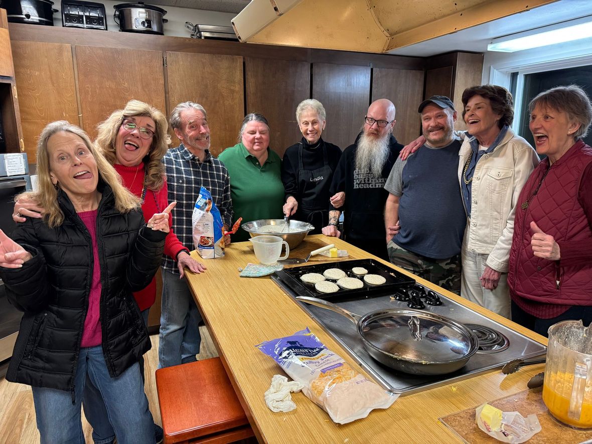Group of people smiling around a kitchen counter with food and drinks during a casual gathering