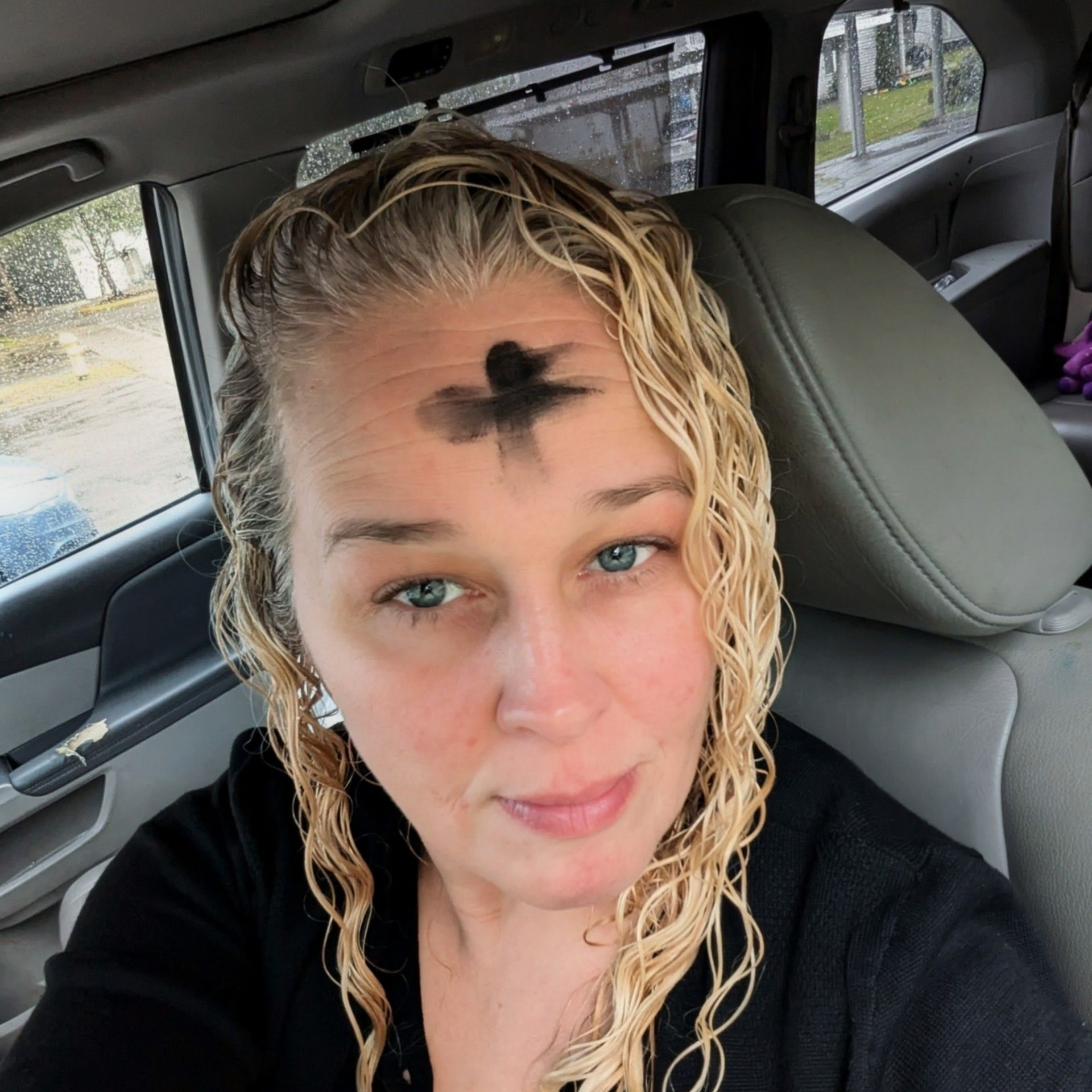 Woman with wet blonde hair sitting in a car, looking at the camera with a black ash cross on her forehead.