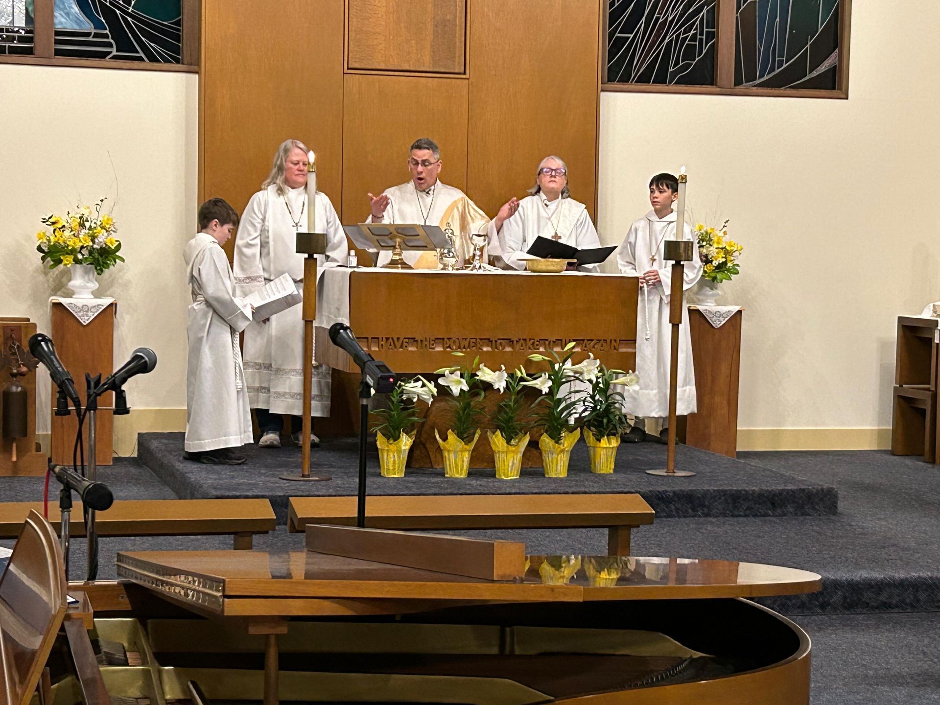 Clergy in white robes stand behind an altar in a church, with flowers and candles on the altar.