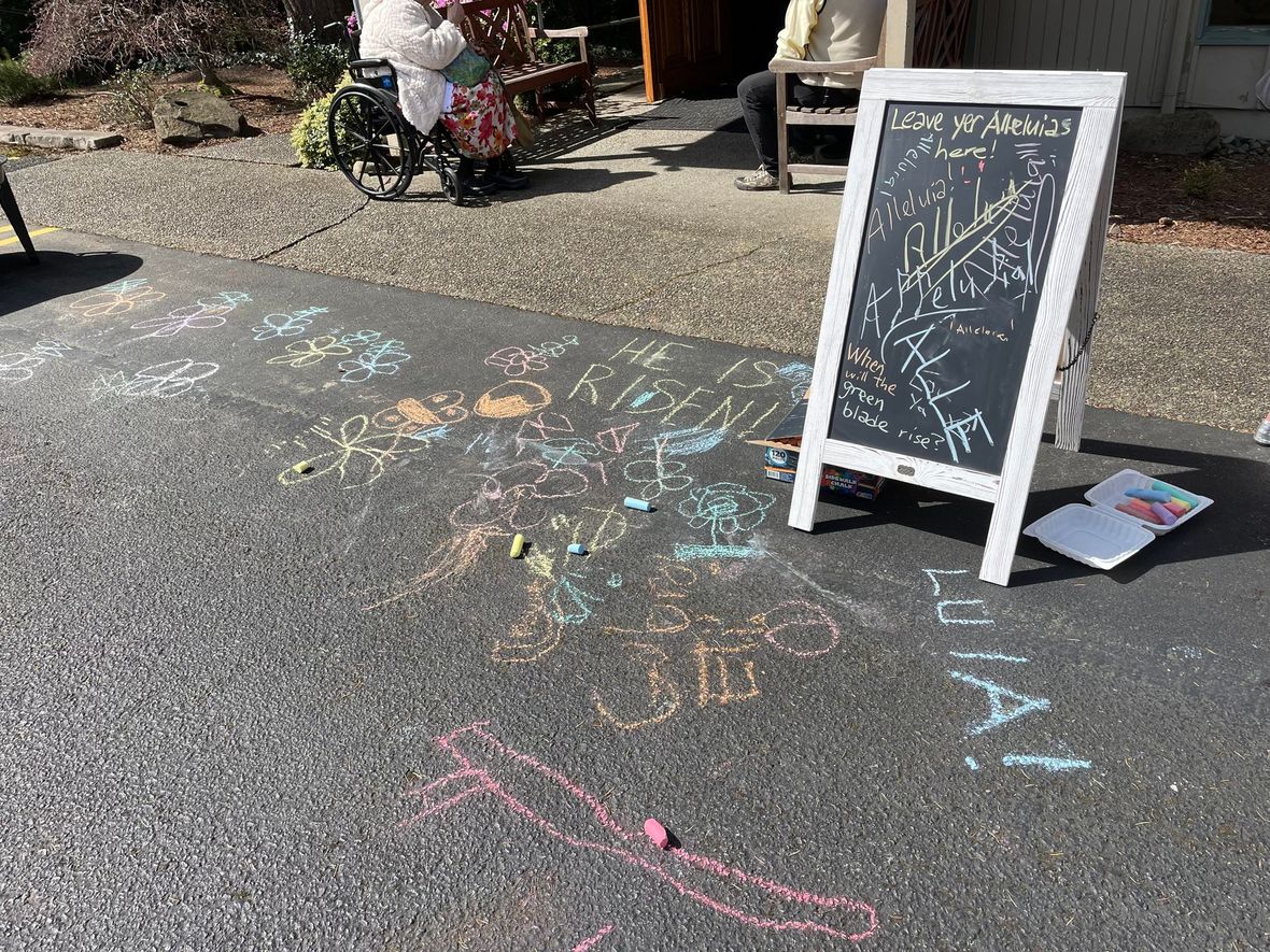 A sidewalk chalkboard sign on an asphalt path with colorful chalk drawings and scribbles nearby