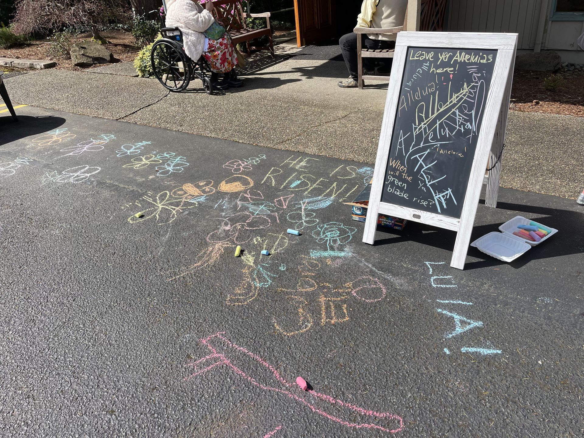 A sidewalk chalkboard sign on an asphalt path with colorful chalk drawings and scribbles nearby