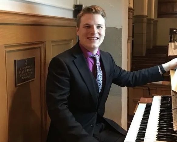 Smiling man in a dark suit seated at a piano in a wood-paneled room