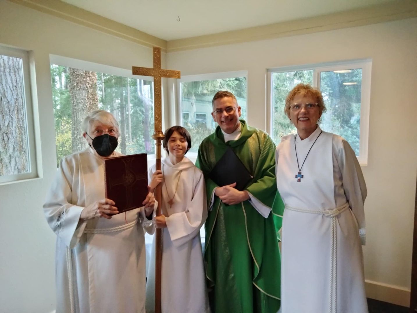 Four clergy in white and green robes stand indoors beside a wooden cross by bright windows.