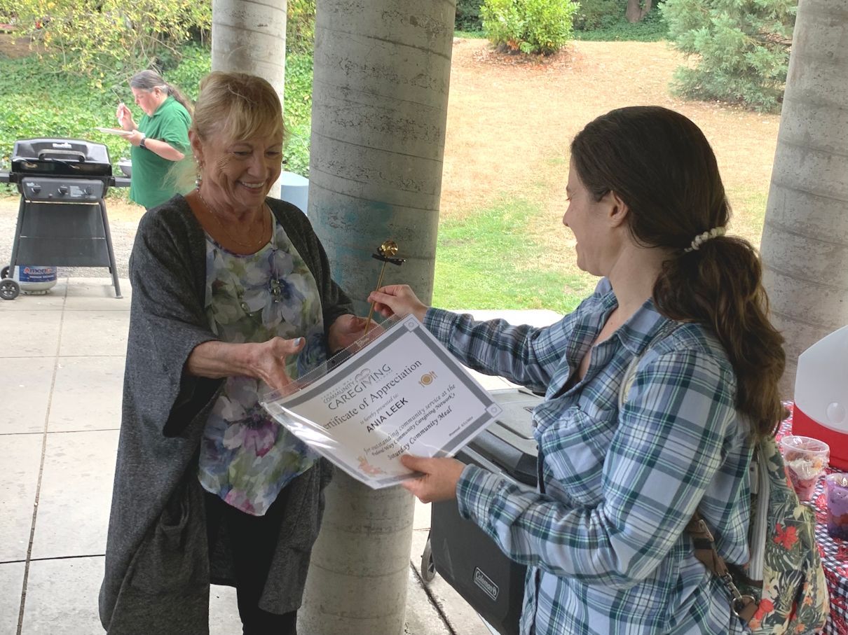 Woman hands a certificate to another person outdoors under a covered area.