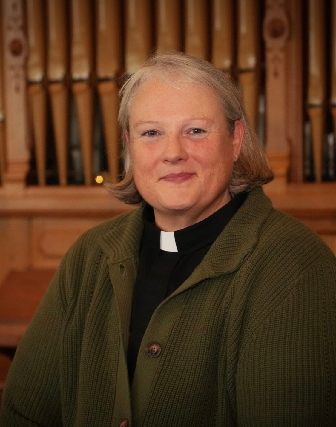 Person in a clerical collar and green cardigan seated in front of wooden organ pipes, smiling.