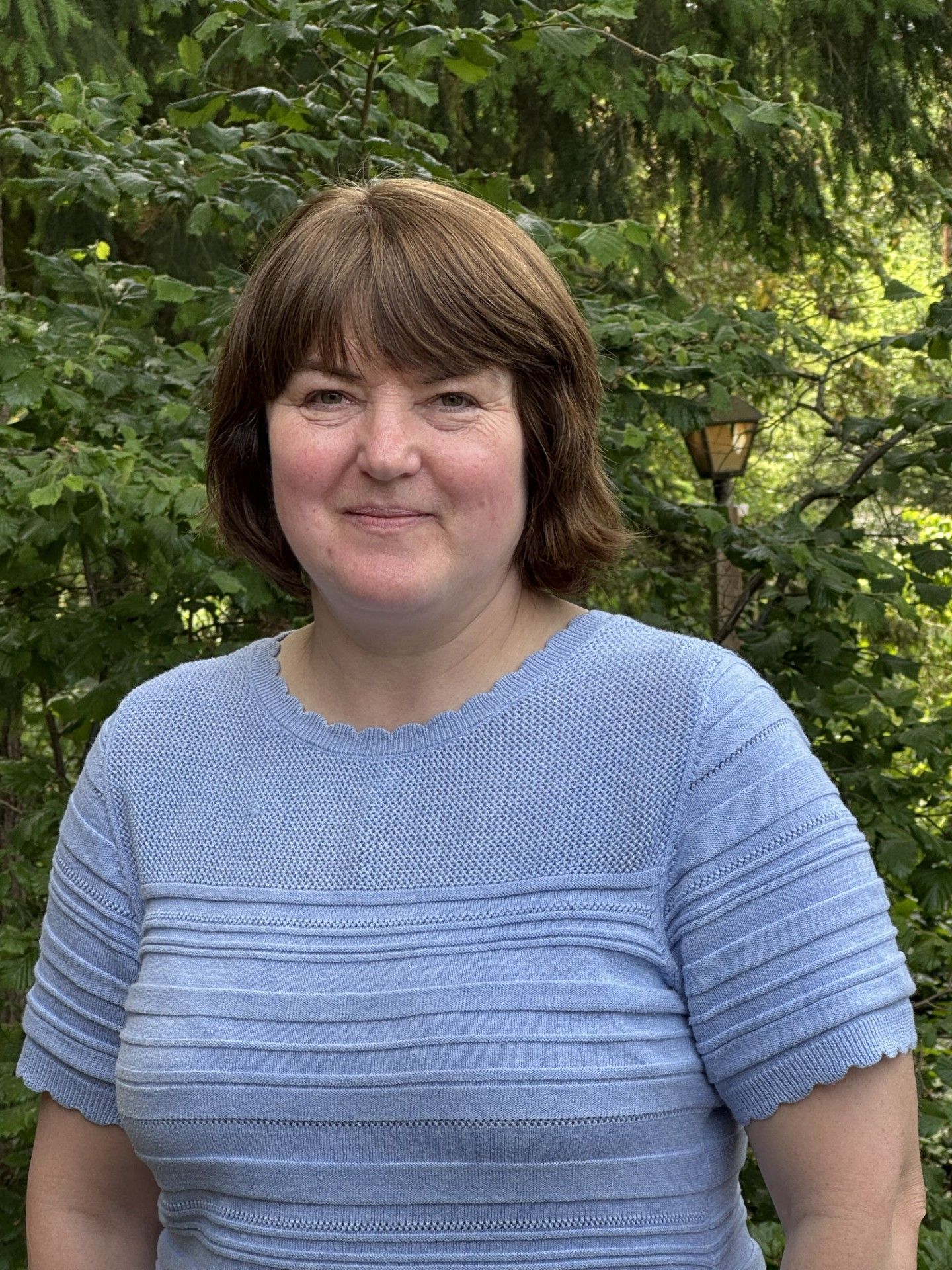 Smiling person in a light blue sweater standing outdoors amid green foliage.