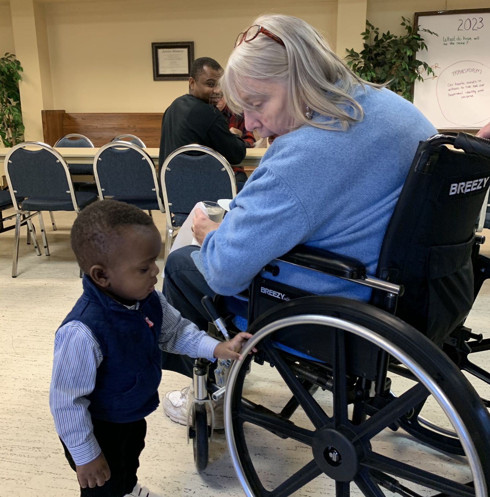 Child standing beside a wheelchair user in a community room, with chairs and a whiteboard in the background
