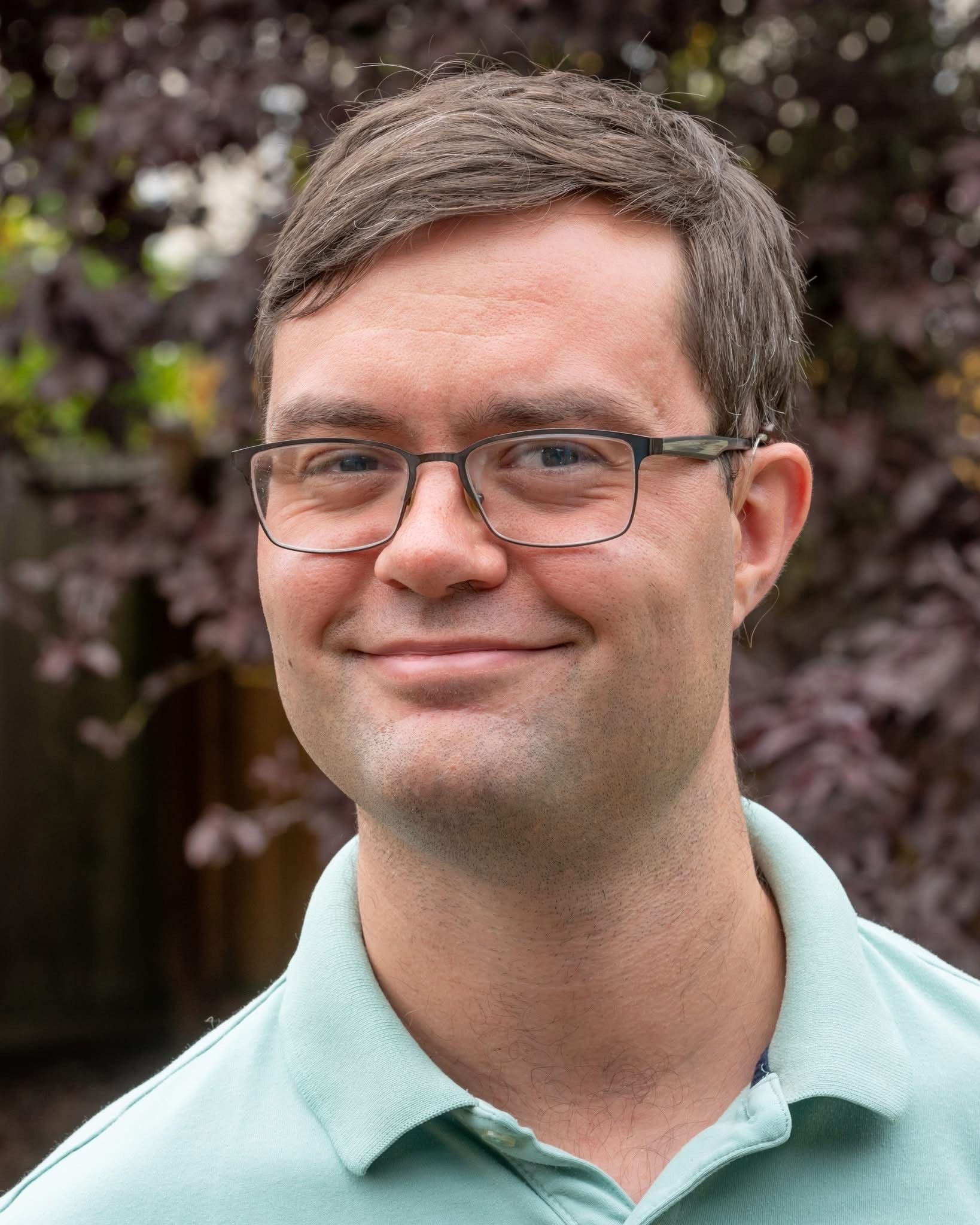 Smiling man with glasses in a light green shirt, standing outdoors against a leafy background