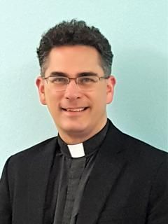 Portrait of a smiling person in a black clerical suit and white collar against a light blue background