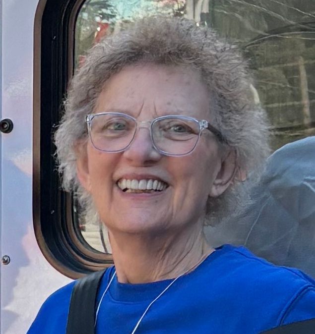 Smiling person with glasses and curly hair wearing a blue shirt outdoors near a vehicle window