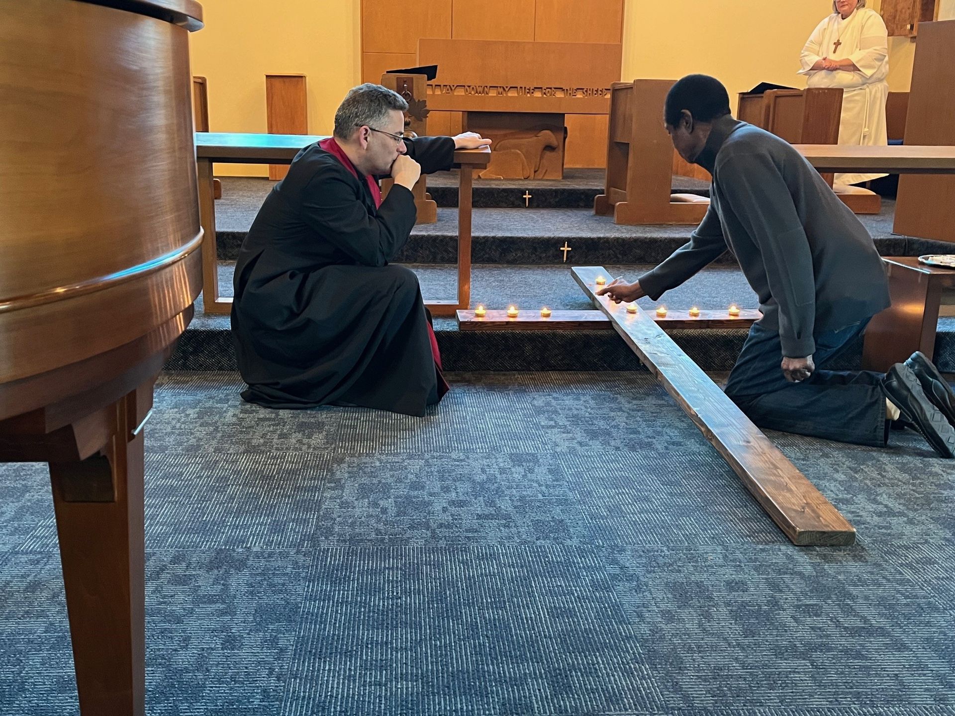 Two people kneel in a church, praying at a wooden cross during a solemn ceremony.