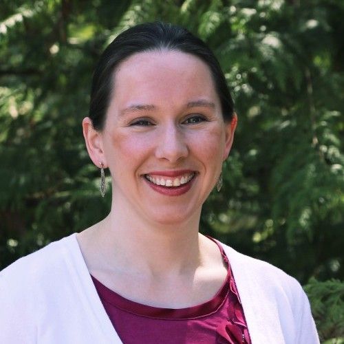 Smiling woman outdoors in a white cardigan and maroon top, with green trees in the background