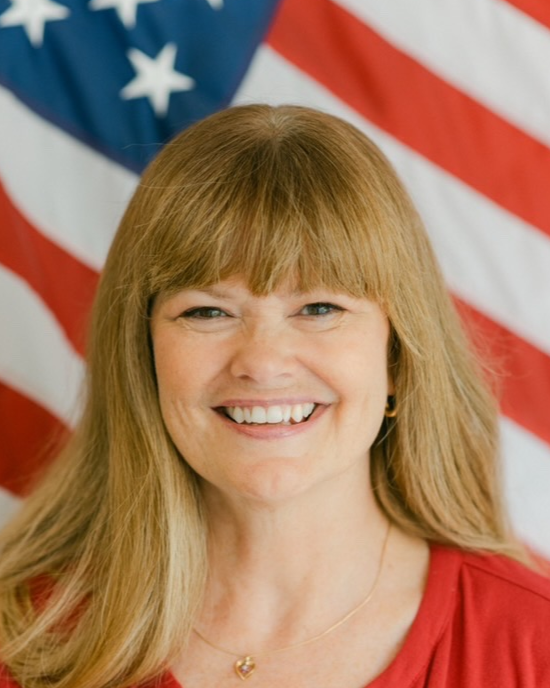 Woman smiling, blond hair, red shirt, in front of American flag.