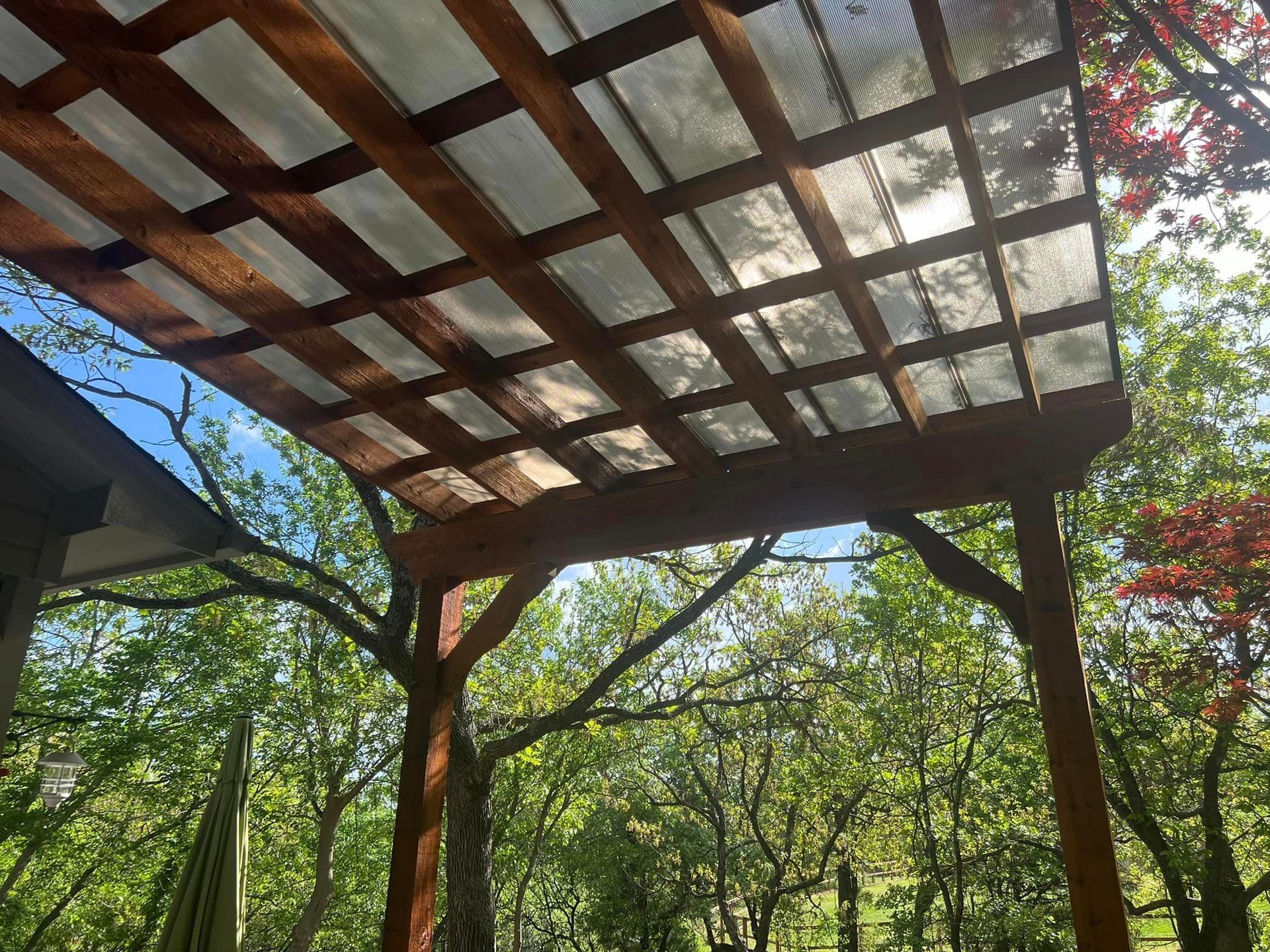 A wooden pergola with a clear plastic roof is surrounded by trees.