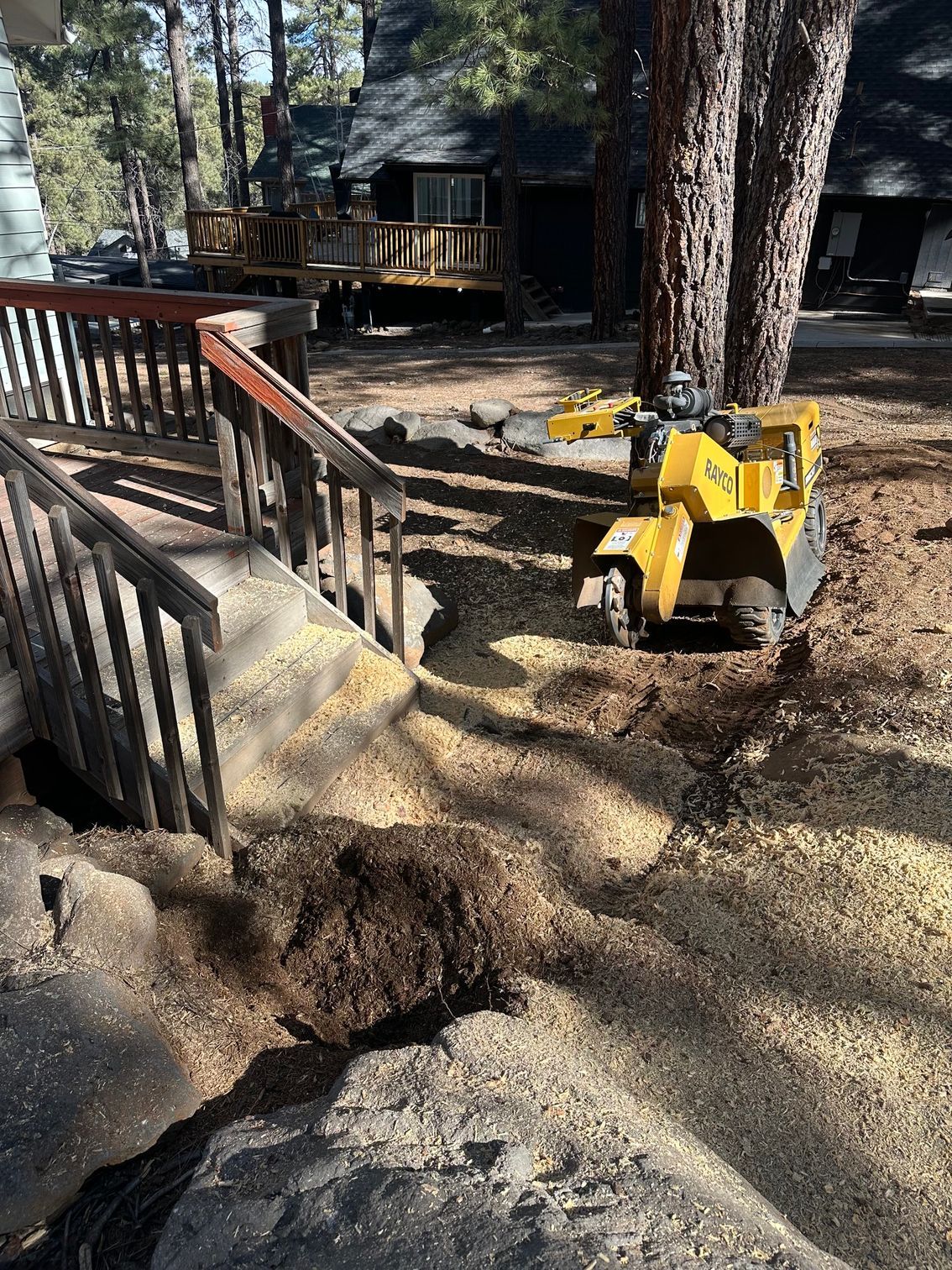 Yellow construction equipment beside a wooden deck and railing in a wooded yard