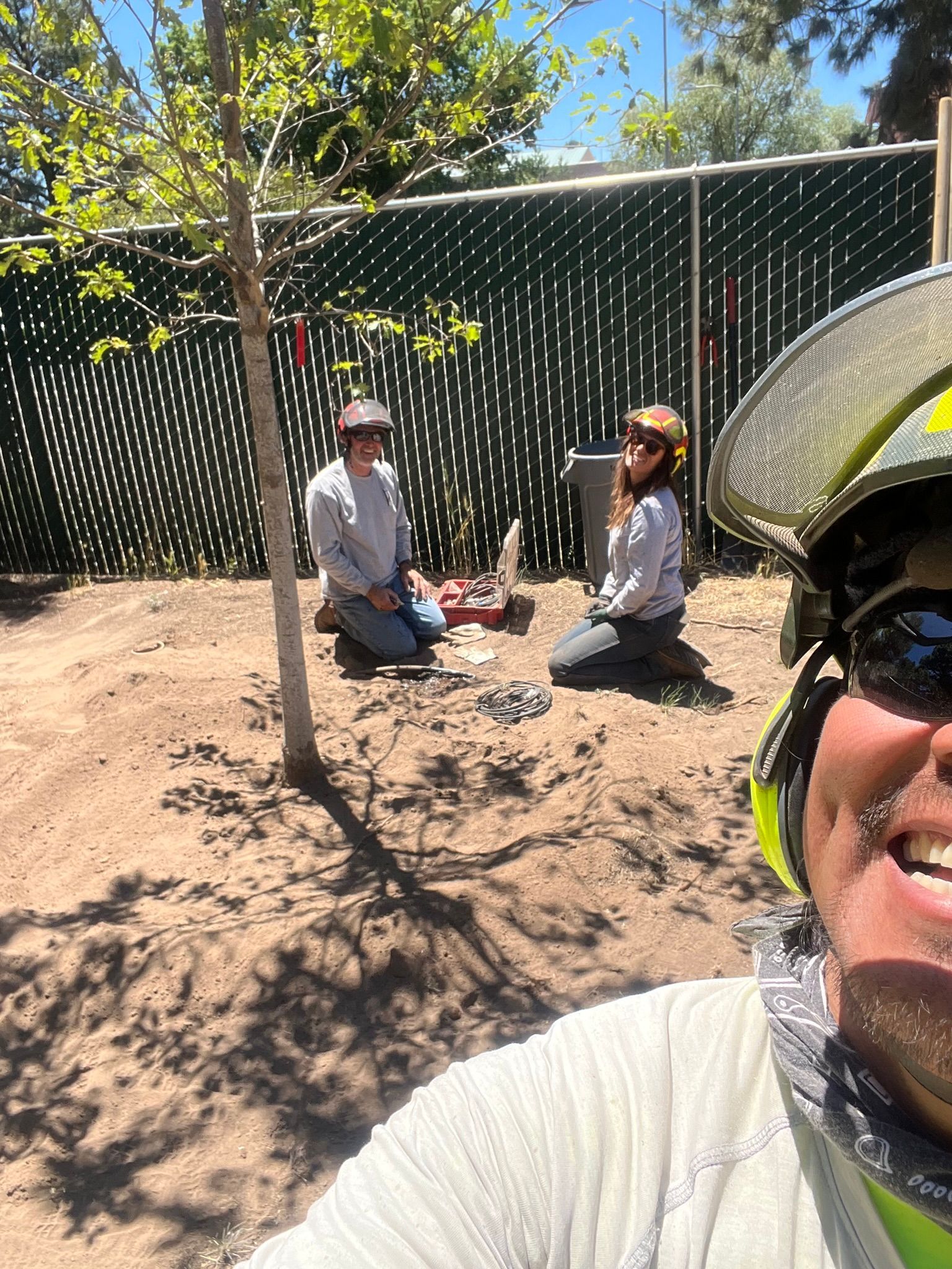 Two people sitting on the ground in a shaded outdoor yard, with a smiling cyclist close to the camera.