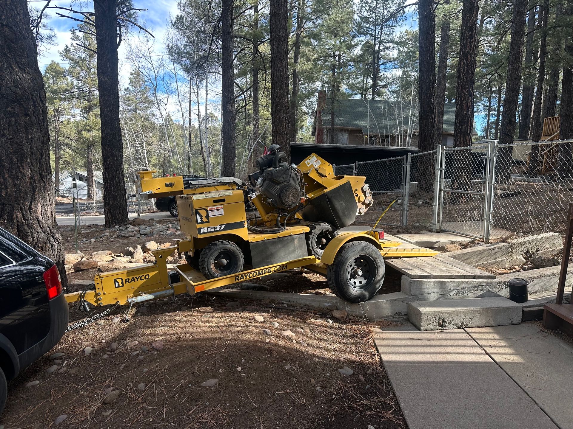Yellow skid steer on a trailer near a driveway, with a black car and trees in the background