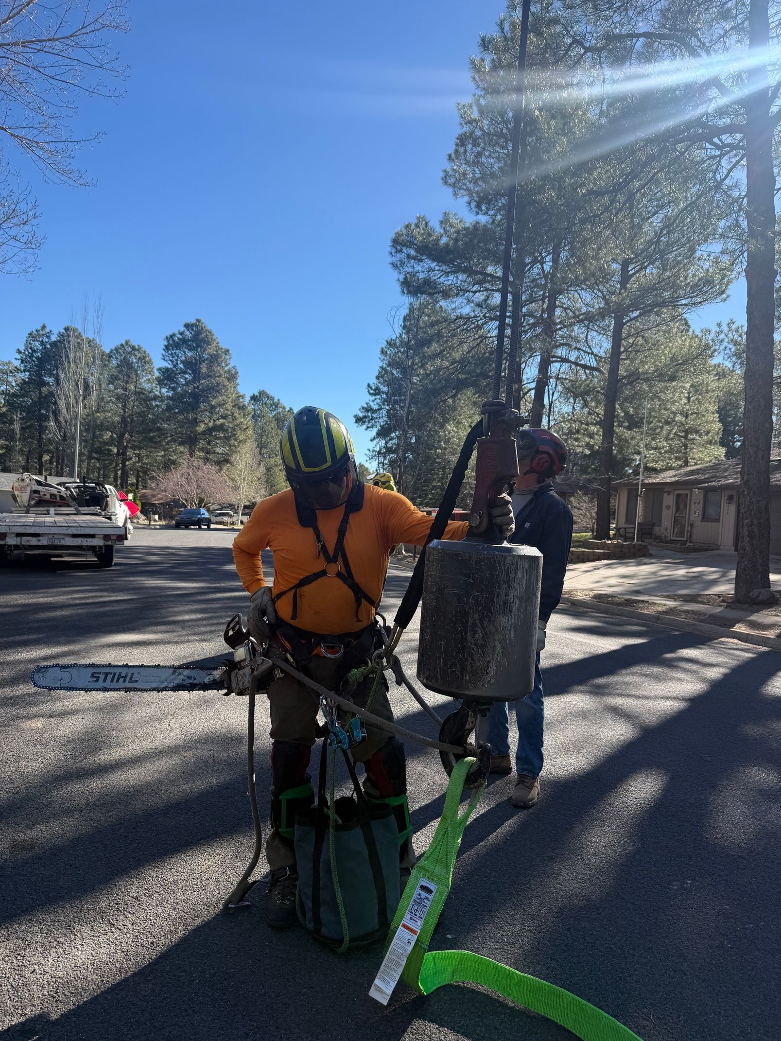 Two utility workers with safety gear stand on a sunny gravel roadside near a truck and trees.