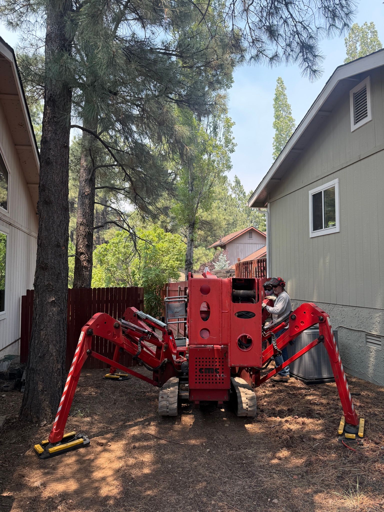 Red spider-like construction robot parked between a house and tall trees in a narrow driveway.