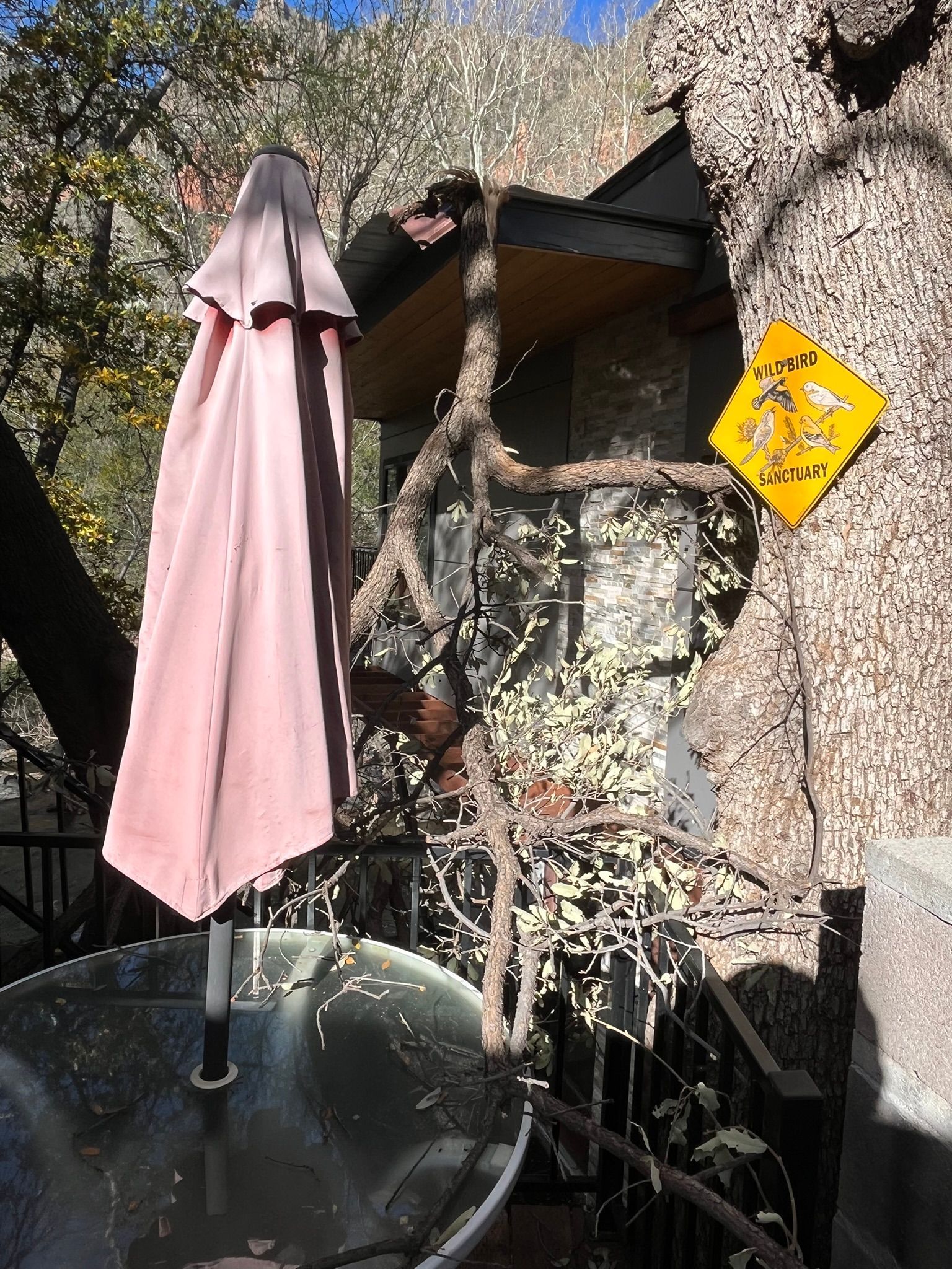 Patio with closed pink umbrella, glass table, stone wall, and yellow sign in sunlight