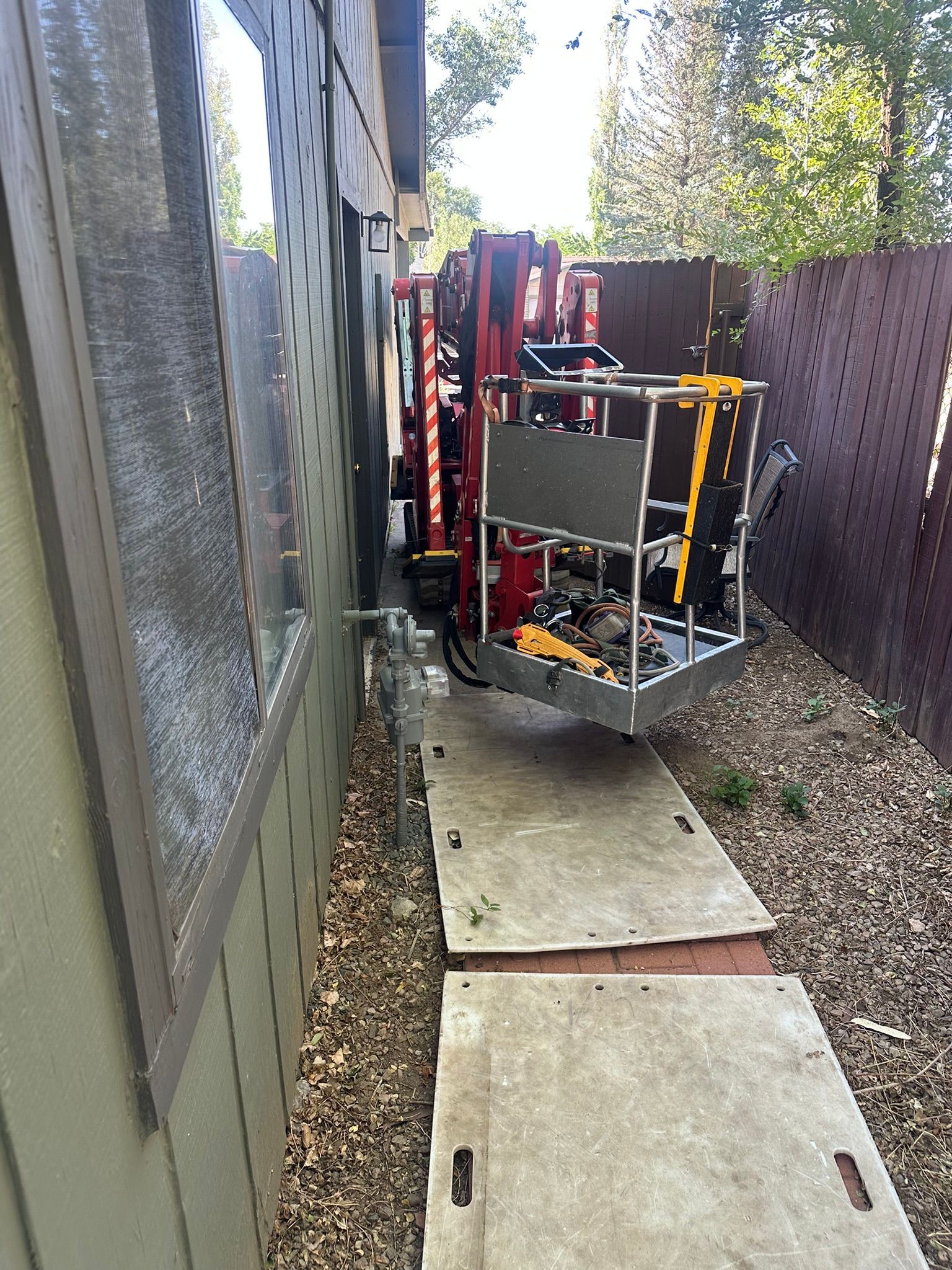 Construction lift in a narrow outdoor path between a house and fence, with wooden planks and debris.