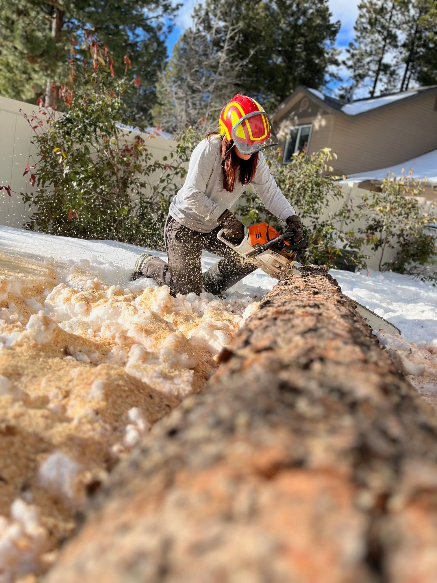 Worker in safety gear chainsaws a fallen tree trunk in the snow near houses.