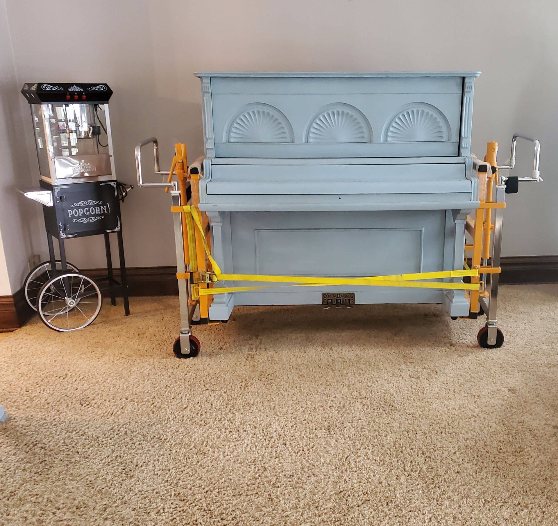 A blue piano on a yellow moving cart and a popcorn machine are inside a room.