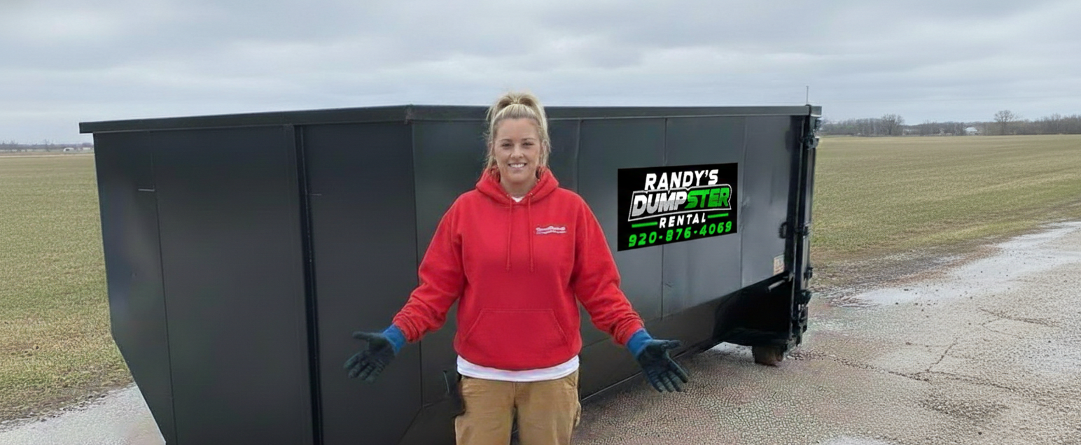 Person in a red jacket stands beside a black dumpster in a muddy field.
